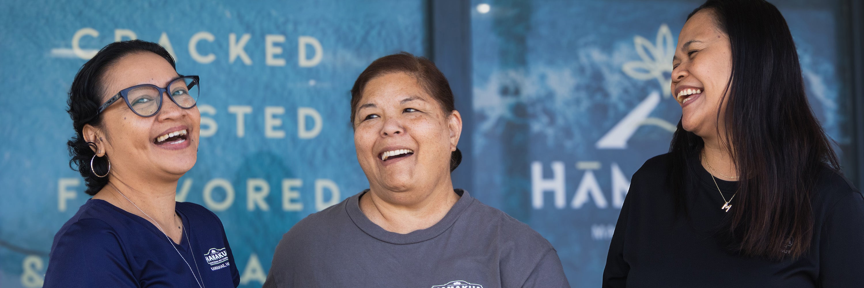 Three Hāmākua Macadamia Nut Company employees laughing together in front of the Visitor Center.