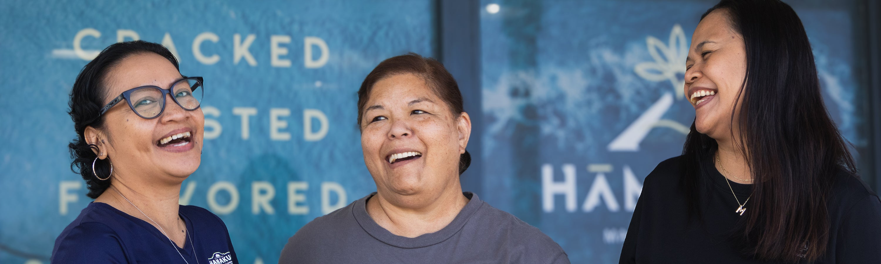 Three Hāmākua Macadamia Nut Company employees laughing together in front of the Visitor Center.