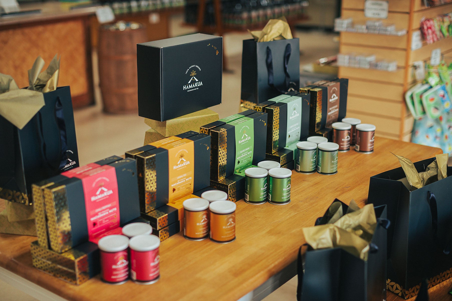 Display of Hāmākua Macadamia Nuc Company packages, boxes, and cans on a wooden table with a store interior in the background.