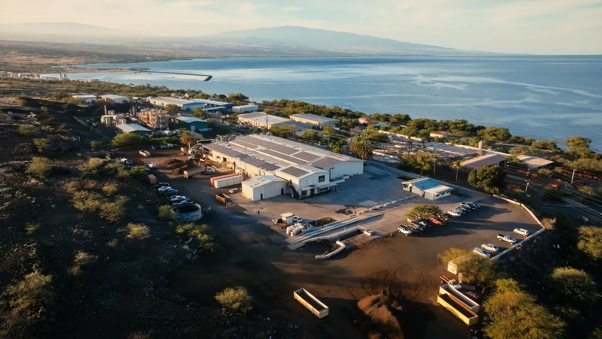 Aerial view of Hāmākua Macadamia Nut Company with white buildings, parking areas, and solar panels, situated near the ocean with mountains in the distance.