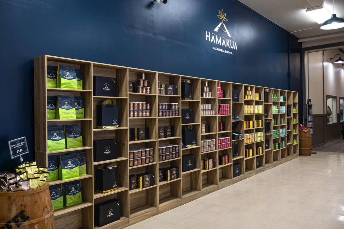 A long wooden shelving unit filled with packaged goods and bags in a retail interior. The wall behind the shelves is dark blue with the Hāmākua Macadamia Nut Co. logo above.